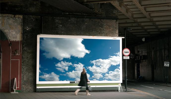 A woman turns to look at a large billboard image of white clouds in a blue sky while walking along a sidewalk.