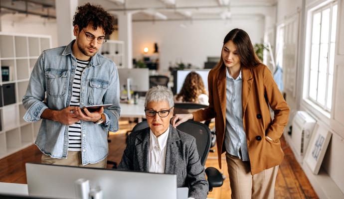 Three casually dressed employees in a modern office meet around a desktop computer.