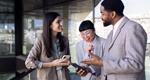 Three colleagues, standing together in an office, smile and laugh while gesturing toward their smartphones
