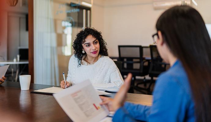 Two colleagues sit at a table in an informal workspace. In the foreground, one of them presents information from a printout, as the other takes notes.