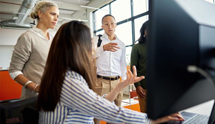 Coworkers meet and gesture toward a computer monitor during a discussion in their office.