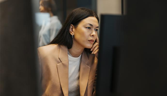 A woman studies a computer monitor with her hand raised to the side of her face. She is framed by additional monitors in the foreground.