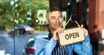 A man wearing an apron peers out of a store window as he holds an “open” sign.