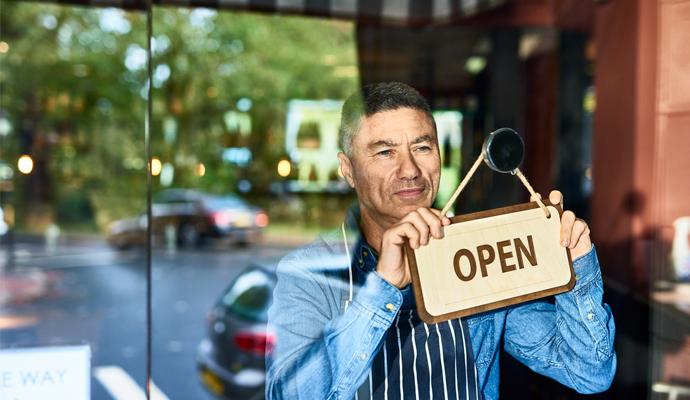 A man wearing an apron peers out of a store window as he holds an “open” sign.