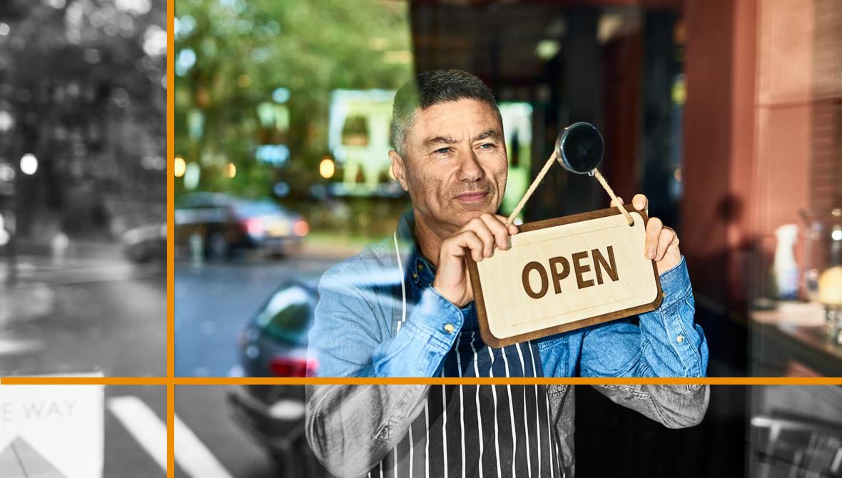 A man wearing an apron peers out of a store window as he holds an “open” sign.