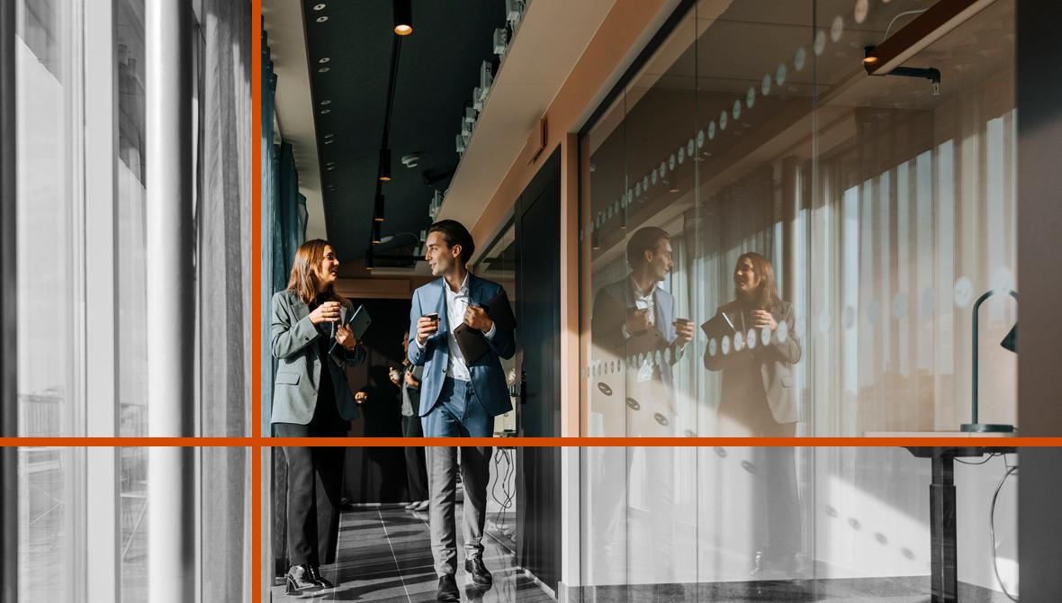 Two colleagues converse as they walk through an office corridor, with coffee cups, laptops, and tablets in hand. The scene is lit through a row of windows that appears to the left, and to the right, the colleagues' reflections are visible in a glass wall.