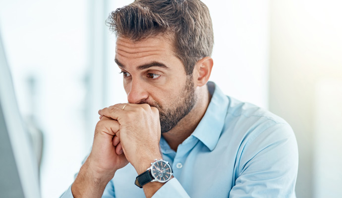 A man sits at a desk with his brow furrowed and his folded hands pressed to his mouth. A man sits at a desk with his brow furrowed and his folded hands pressed to his mouth.