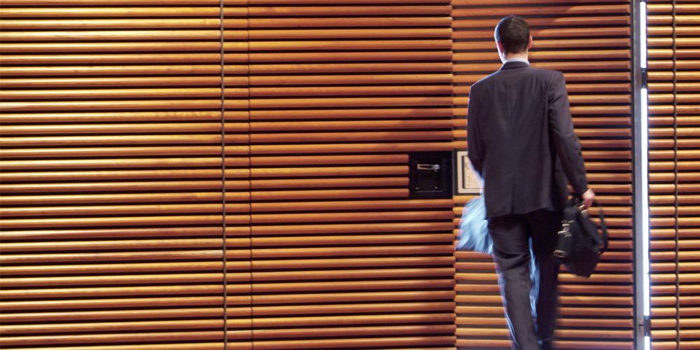 A businessman is seen from behind as he exits the door of a wood-paneled office. 