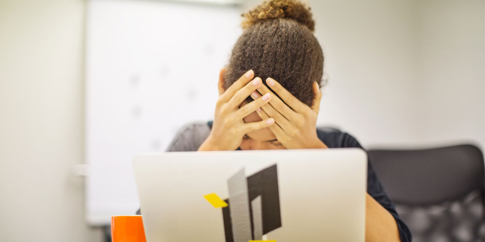 A woman with her head in her hands is partially obscured by an open laptop computer in front of her. A woman with her head in her hands is partially obscured by an open laptop computer in front of her.
