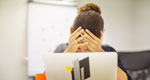 A woman with her head in her hands is partially obscured by an open laptop computer in front of her.