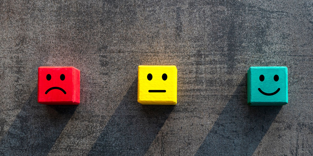 Three colored wooden blocks are next to one another, representing customer satisfaction levels: a red block has a frowning face, a yellow block has a neutral face, and a green block has a smiling face.