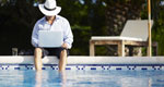 A businessman with a drink and a laptop dangles his feet into a swimming pool.