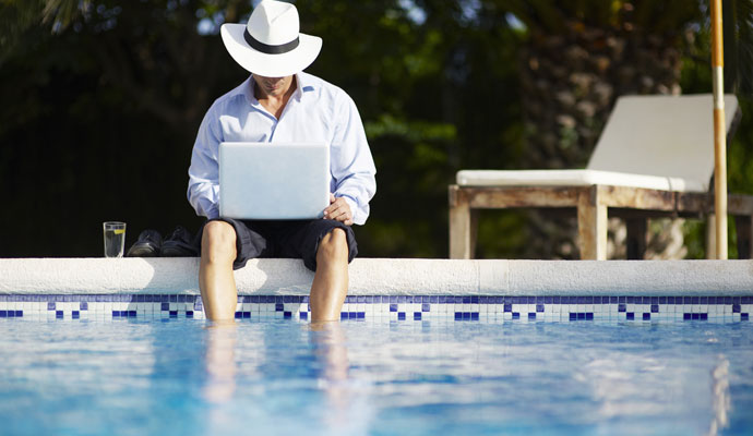 A businessman with a drink and a laptop dangles his feet into a swimming pool. A businessman with a drink and a laptop dangles his feet into a swimming pool.