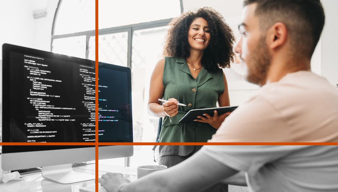 Coworkers collaborate to develop code: a man types at a computer in the foreground as a woman holds a tablet and looks on.