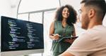 Coworkers collaborate to develop code: a man types at a computer in the foreground as a woman holds a tablet and looks on.