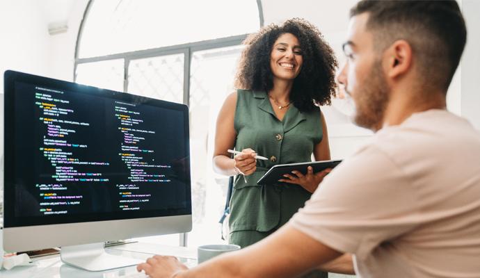 Coworkers collaborate to develop code: a man types at a computer in the foreground as a woman holds a tablet and looks on.