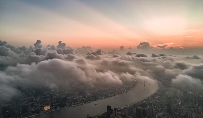 A river winds through a darkened cityscape, with rays of sun emerging from behind a cluster of low clouds. A river winds through a darkened cityscape, with rays of sun emerging from behind a cluster of low clouds.