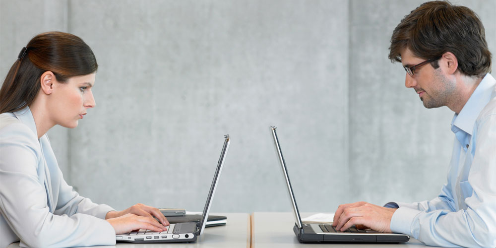 Two businesspeople at laptops, facing each other across a table, as they would in a chess match Two businesspeople at laptops, facing each other across a table, as they would in a chess match