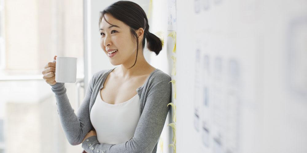 A young woman holding a coffee mug leans against a whiteboard. A young woman holding a coffee mug leans against a whiteboard.