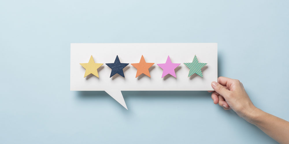 A hand holds a cutout of a white speech bubble against a light-blue background. In the speech bubble are five stars, each a different color. A hand holds a cutout of a white speech bubble against a light-blue background. In the speech bubble are five stars, each a different color.