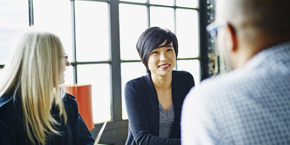Three coworkers sit at a conference room table, talking and smiling.