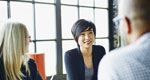 Three coworkers sit at a conference room table, talking and smiling.