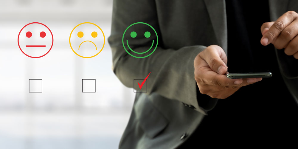 Torso and hands of a man using a smartphone. To his left is an overlay of the image on his screen: a rating system for customer service with a frowning face, a neutral face, and a smiling face. The smiling face is selected. Torso and hands of a man using a smartphone. To his left is an overlay of the image on his screen: a rating system for customer service with a frowning face, a neutral face, and a smiling face. The smiling face is selected.