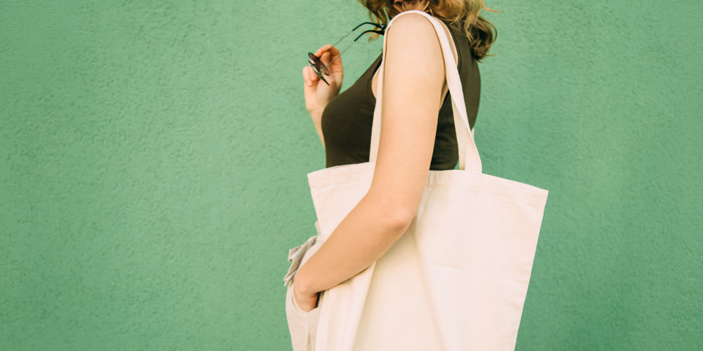 A photo of a woman standing in profile against a green wall, with a cream-colored canvas shopping bag over her shoulder.