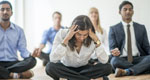 Amid a group of office workers meditating together, a woman is stressed and holds her head in her hands.