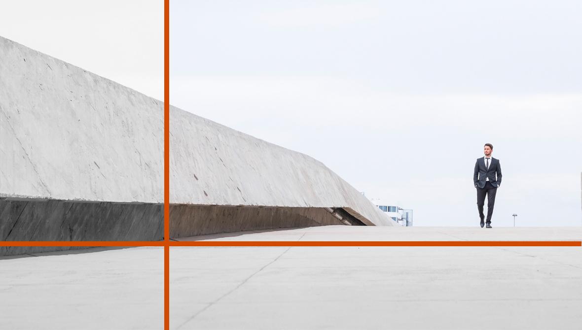 A man in a business suit walks on an empty paved path. Alongside a large concrete boundary and beneath a gray, overcast sky, he appears small and solitary.