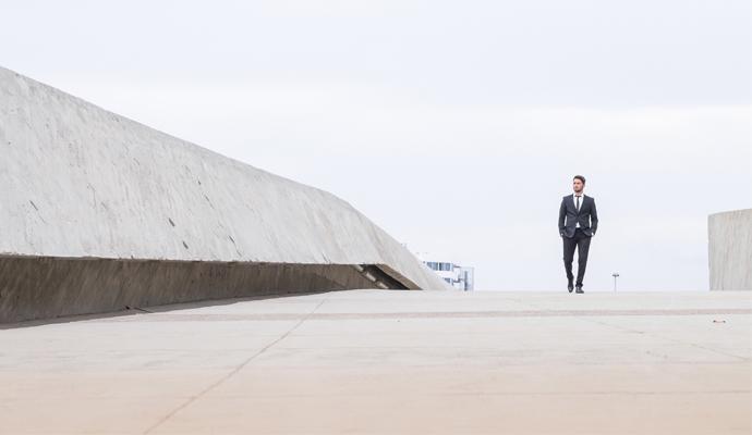 A man in a business suit walks on an empty paved path. Alongside a large concrete boundary and beneath a gray, overcast sky, he appears small and solitary.