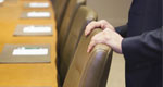 A pair of hands rests on one of the chairs in an otherwise empty boardroom.