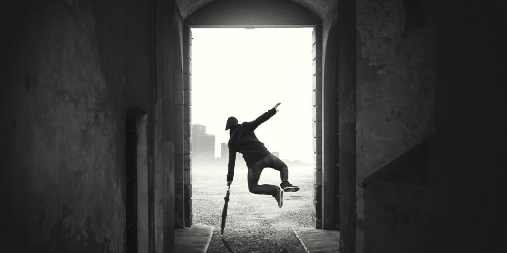 A photograph of a man with an umbrella kicking his heels together on a rainy day.