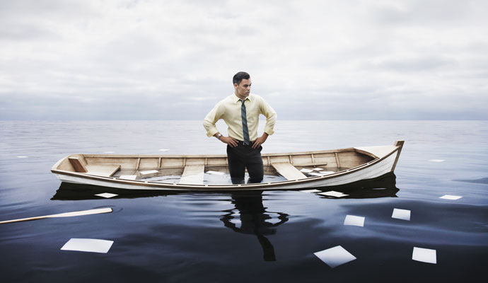 A businessman stands in a sinking boat with oars and papers floating away into an overcast sea
