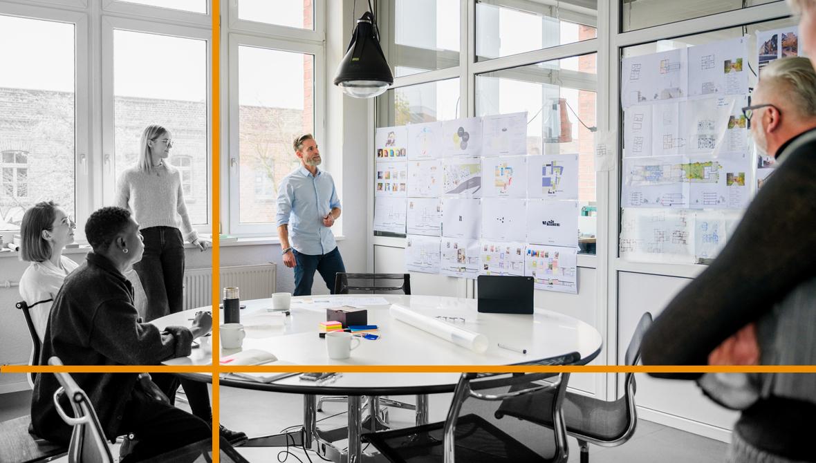 A team of colleagues meets in a small room to discuss data presented on one of the room's walls.