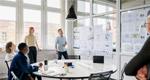 A team of colleagues meets in a small room to discuss data presented on one of the room's walls.
