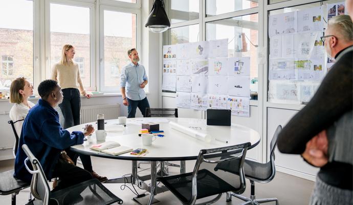 A team of colleagues meets in a small room to discuss data presented on one of the room's walls.