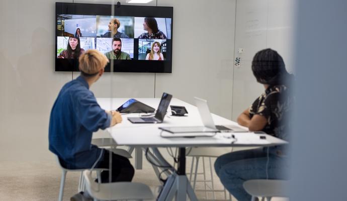 Two people sit in a conference room, with their heads turned toward a screen showing six remote coworkers.