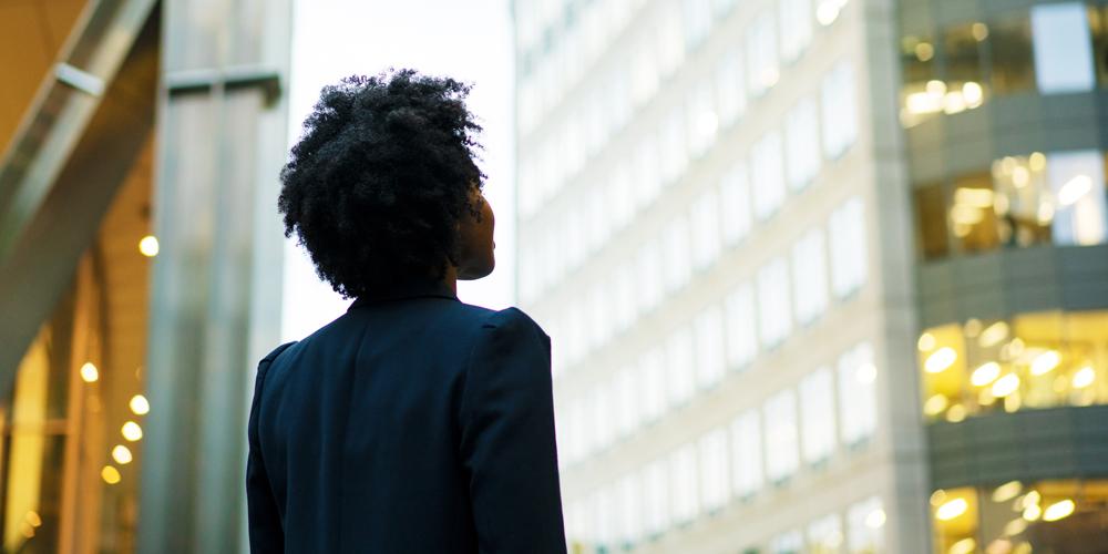 A photograph of a woman in business clothes looking up at an office building. A photograph of a woman in business clothes looking up at an office building.