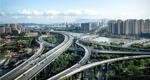Elevated highways pass above trees and cross a river, en route to a city skyline. A mountain range rises in the far distance.