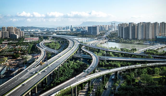 Elevated highways pass above trees and cross a river, en route to a city skyline. A mountain range rises in the far distance.