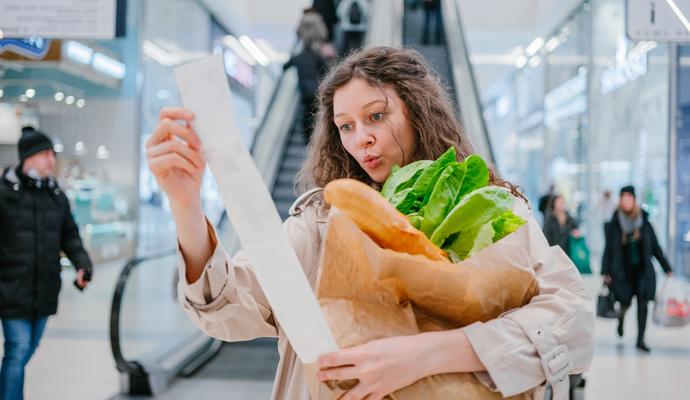 Woman holding a bag of groceries and reviewing her receipt with a surprised look on her face