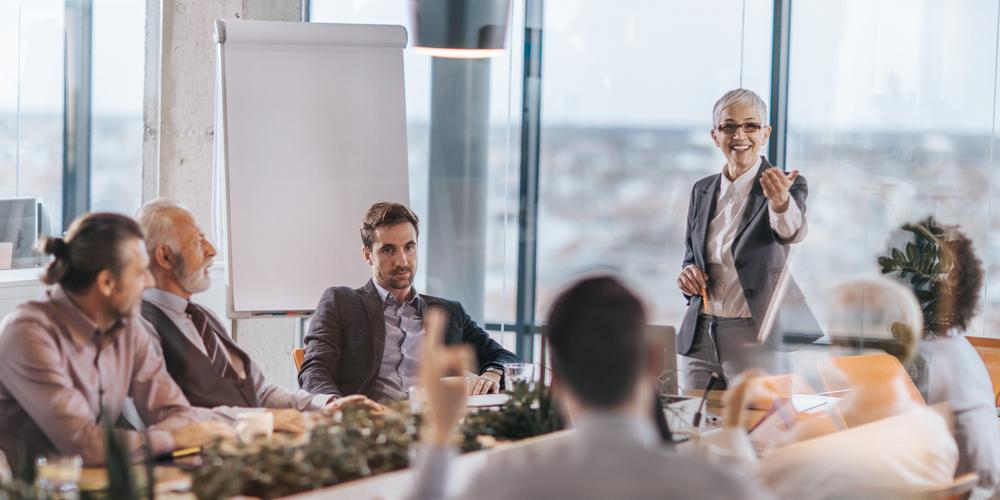 A photograph of a smiling woman in a business meeting gesturing to a man who has his hand raised
