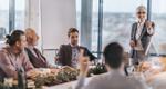 A photograph of a smiling woman in a business meeting gesturing to a man who has his hand raised