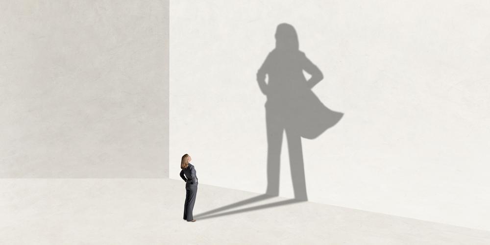 A businesswoman looks at her shadow, which depicts her as a confident, caped superhero. A businesswoman looks at her shadow, which depicts her as a confident, caped superhero.