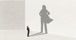 A businesswoman looks at her shadow, which depicts her as a confident, caped superhero.