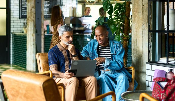 A businessperson has an informal meeting with a client. They're seated together in a cafe as they discuss information presented on a laptop.