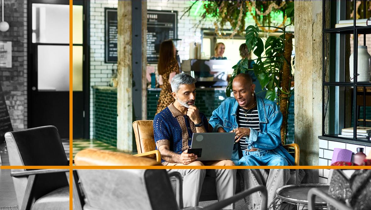 A businessperson has an informal meeting with a client. They're seated together in a cafe as they discuss information presented on a laptop.