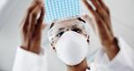 A photograph of a masked medical worker handling a tray of lab samples.