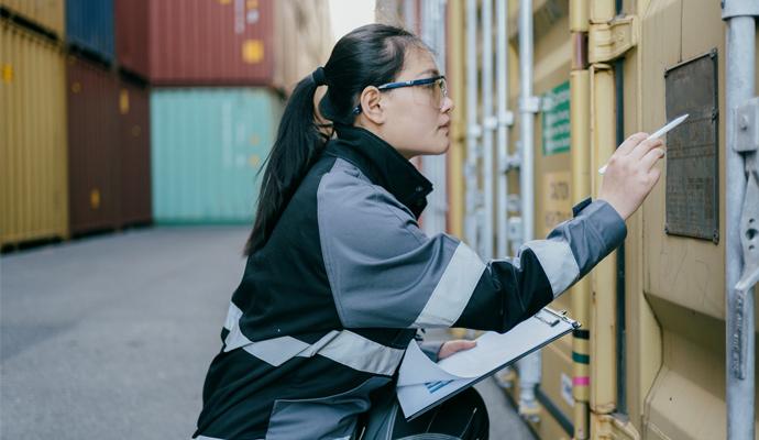 A photo of a woman with a clipboard who is reading the label on a shipping container